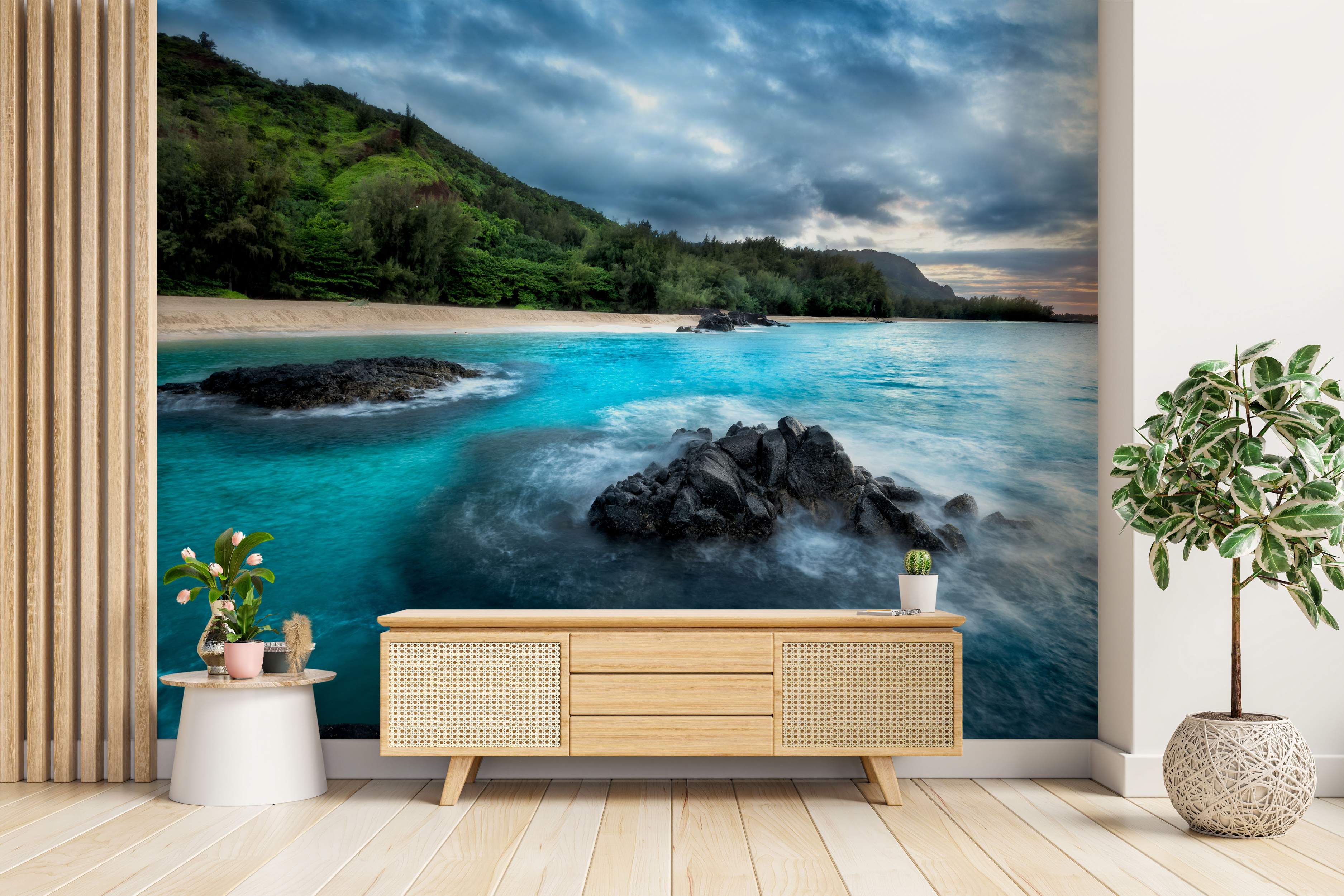Photographic mural depicting a tropical coast with green hills, turquoise water, dark rocks, sandy beach, and dramatic cloudy sky.