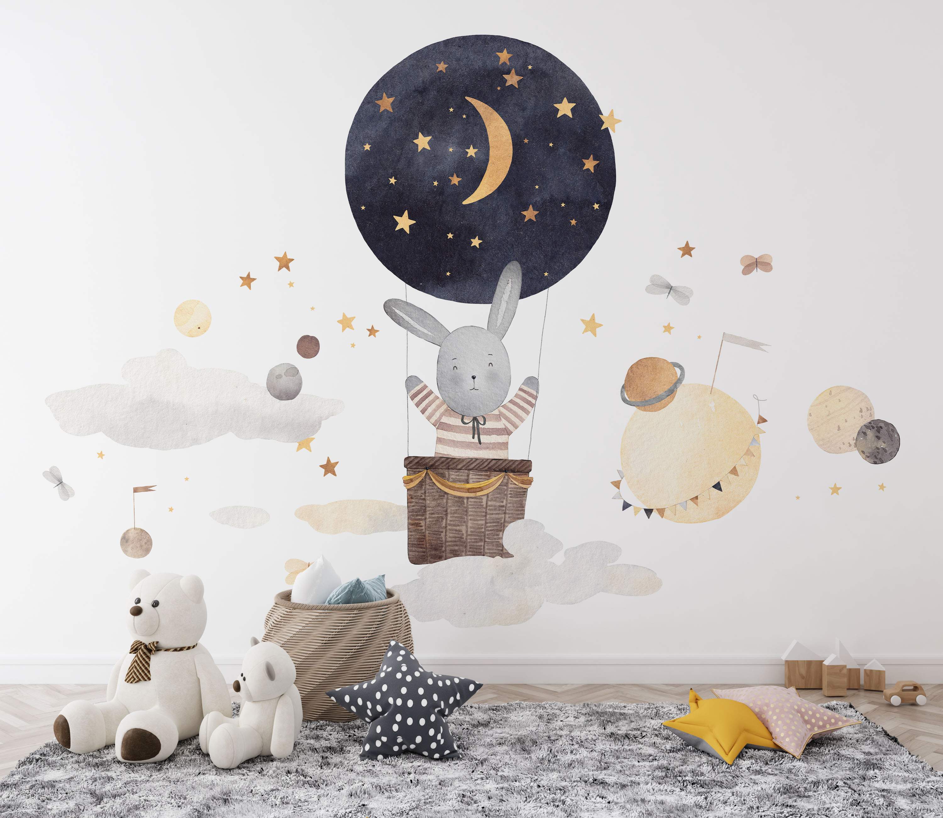 Watercolor mural featuring a gray bunny in a starry blue hot air balloon among clouds, planets, and gold stars.