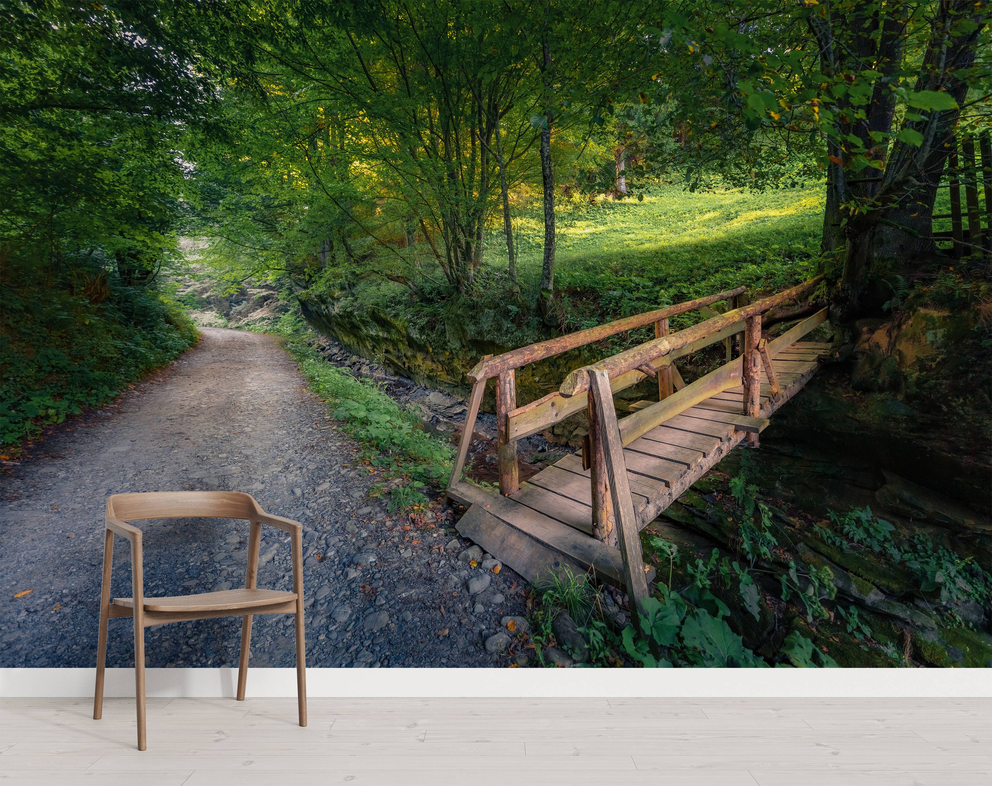 A wall mural of a rustic wooden bridge over a creek next to a dirt path in a lush, sunlit green forest.