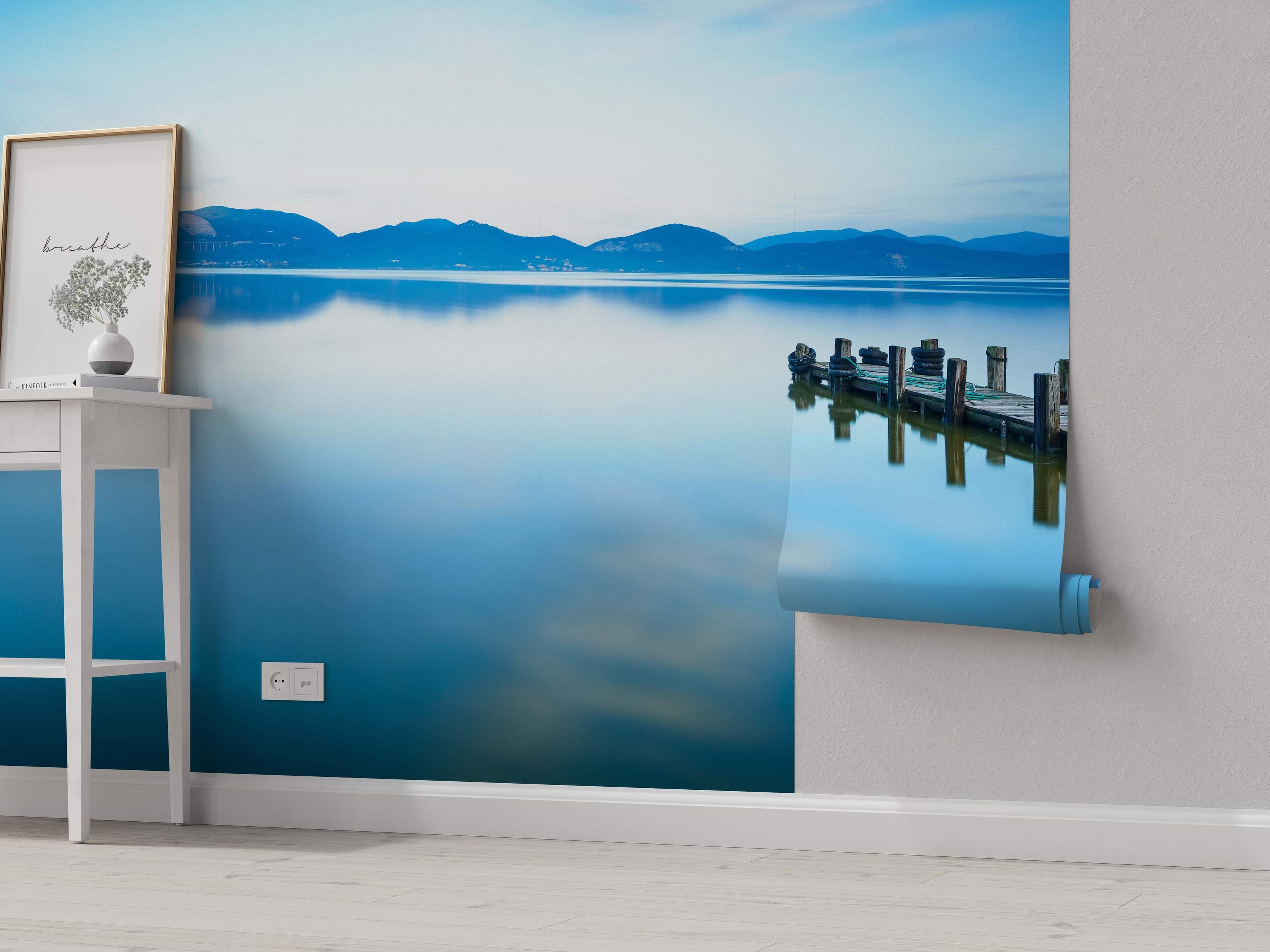 Tranquil lake mural depicting distant blue mountains reflecting in calm water with a wooden dock on the right.