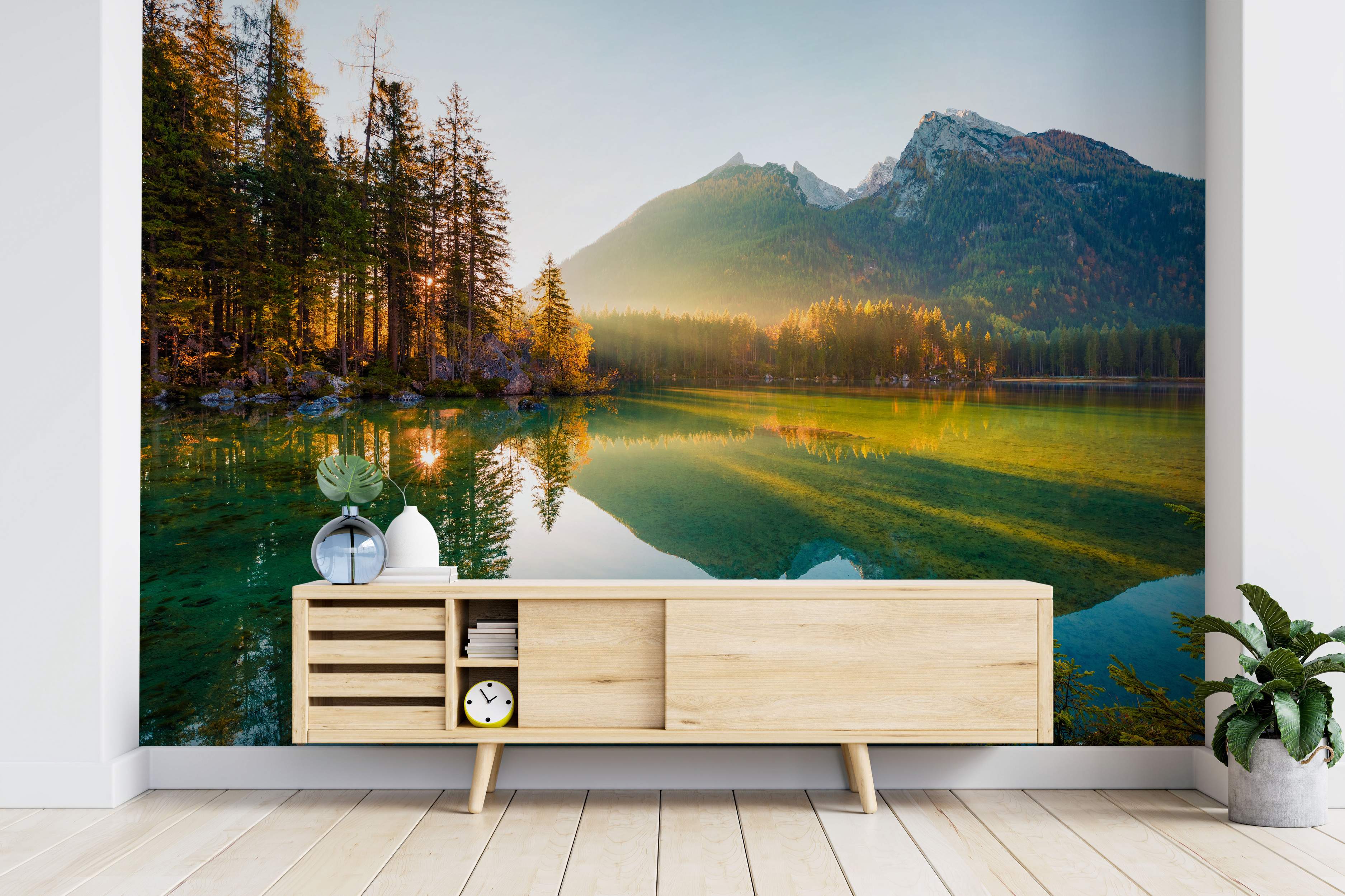 Scenic wall mural depicting a calm turquoise lake reflecting golden sunrise light, evergreen trees, and distant mountains.