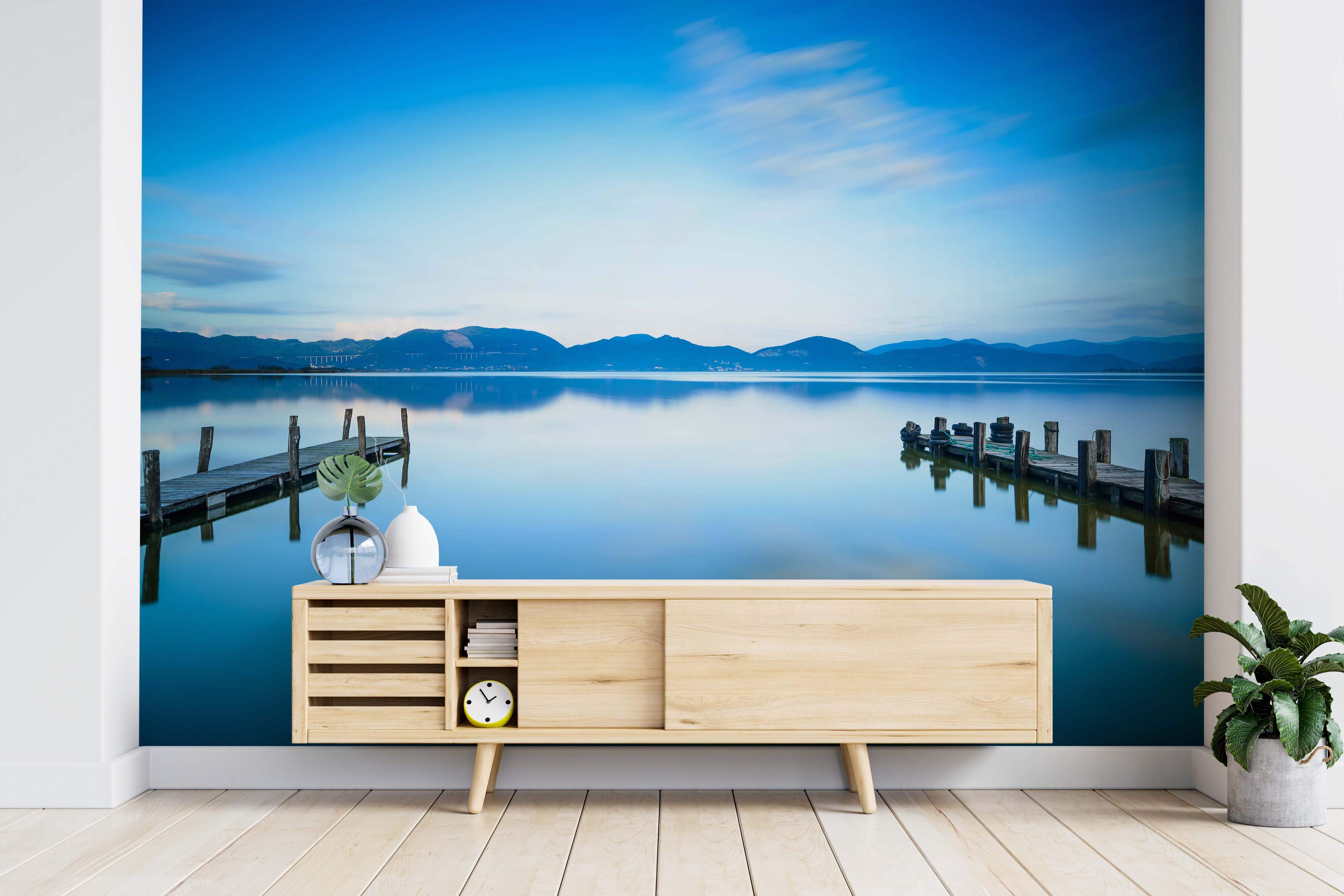 Mural featuring a calm blue lake with two wooden piers reflecting in still water under a blue sky and distant mountains.