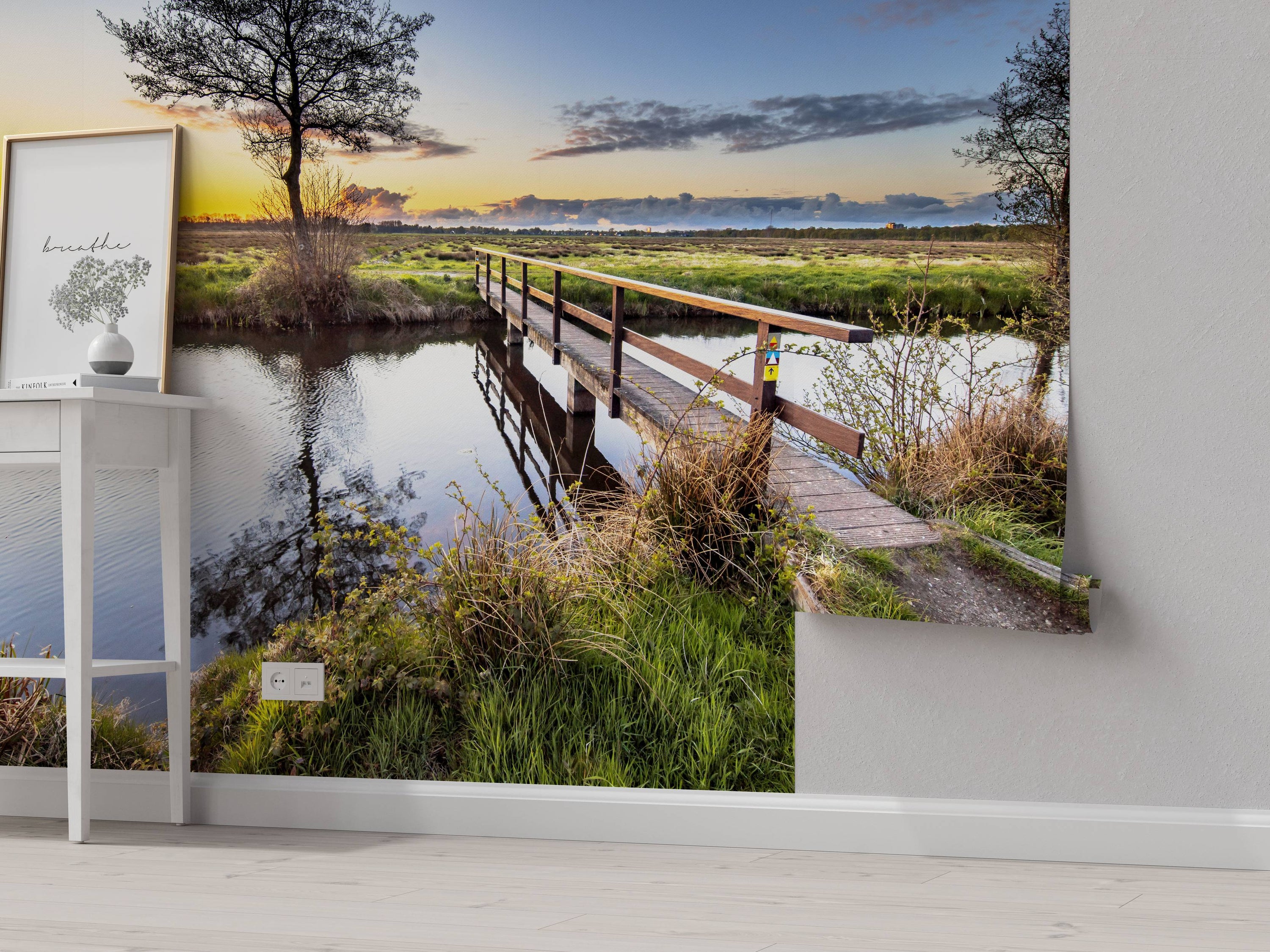 A scenic wall mural of a wooden bridge over a calm river in a grassy field during a colorful sunset.