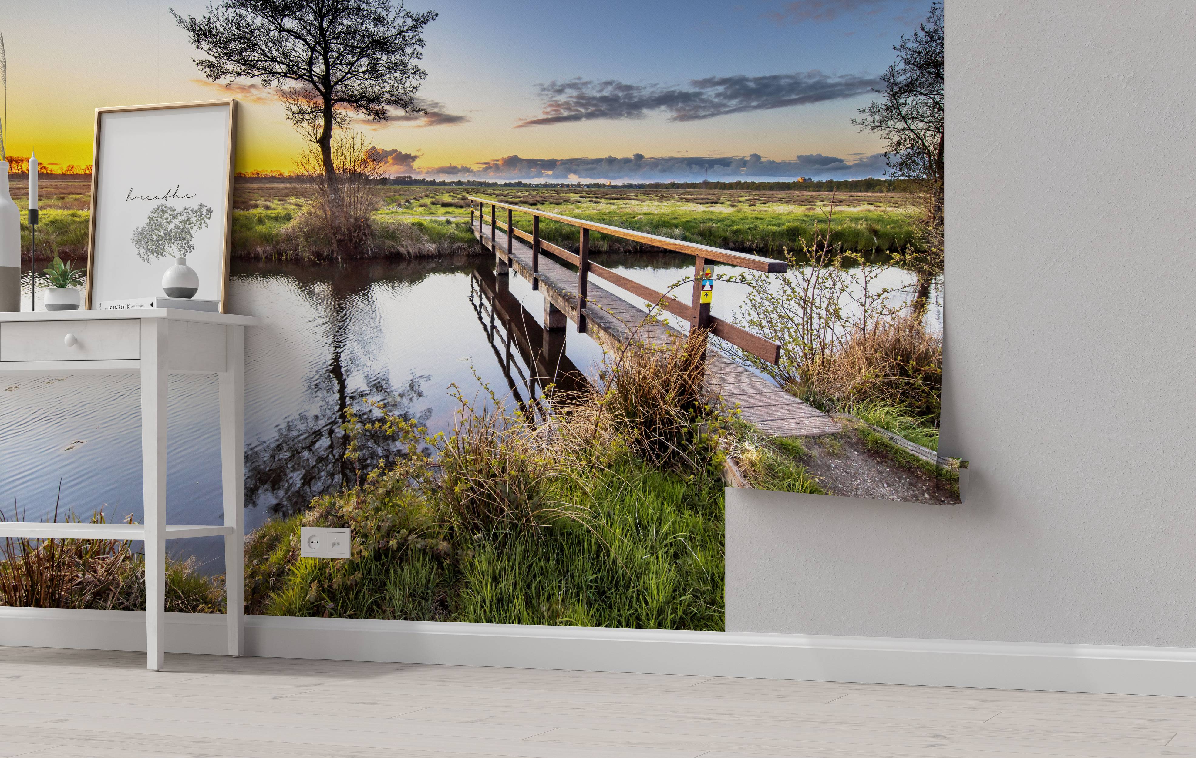 A scenic wall mural of a wooden bridge over a calm river in a grassy field during a colorful sunset.