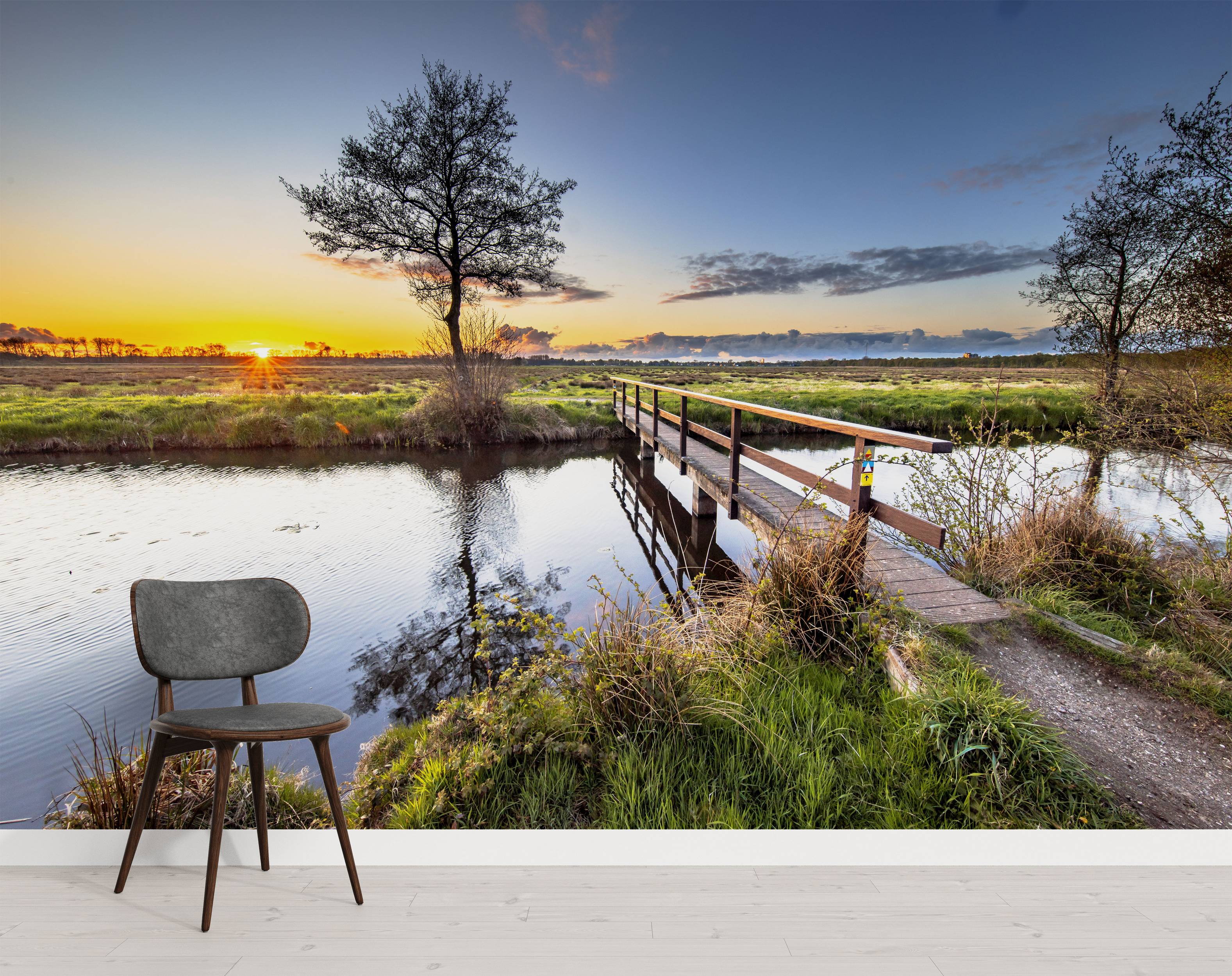 Mural of a scenic sunset over a tranquil river. A wooden bridge crosses the water, with trees reflected in the stream.
