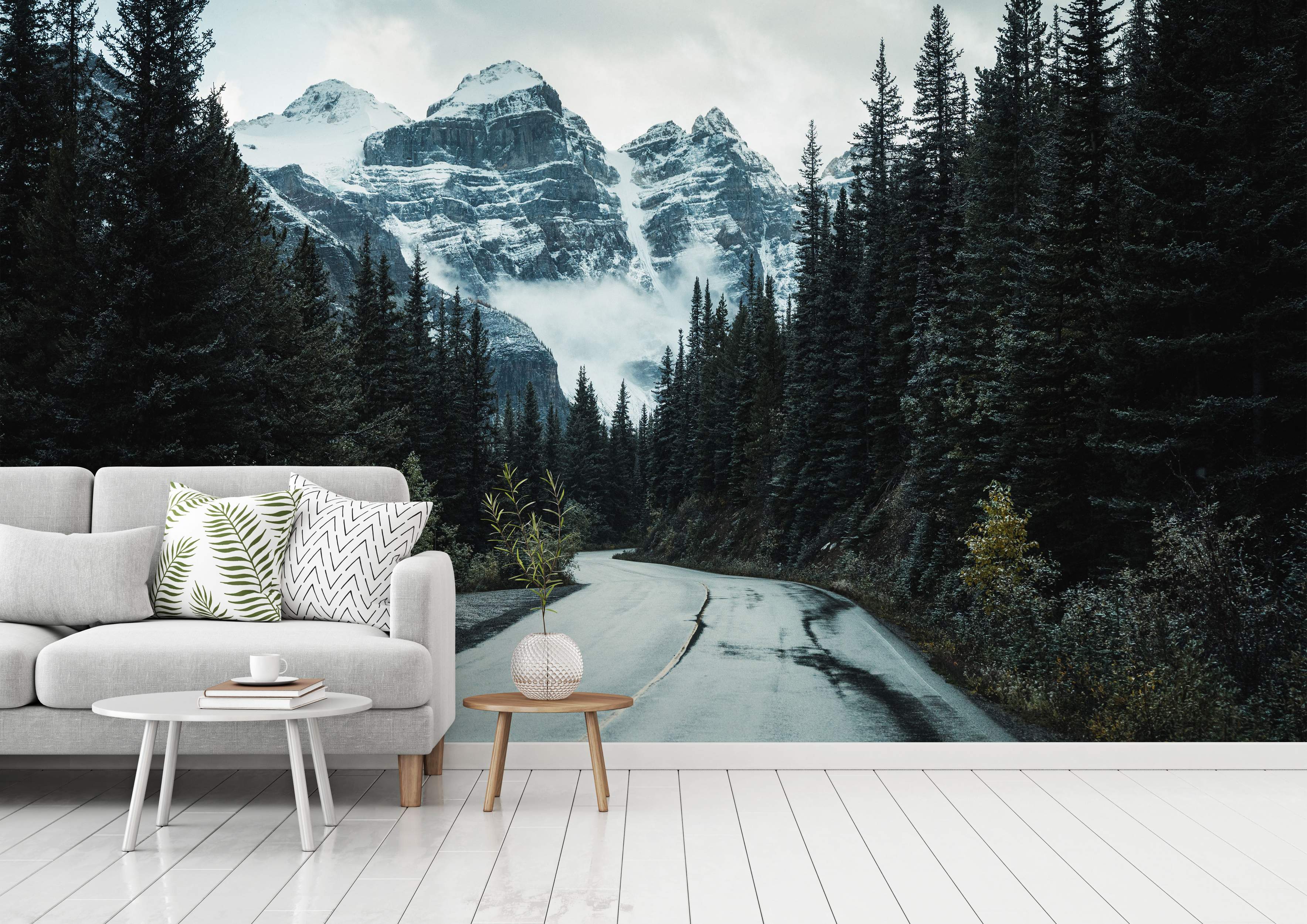 Mural featuring a winding road through a dark green forest towards imposing, snow-capped gray mountains under a cloudy sky.