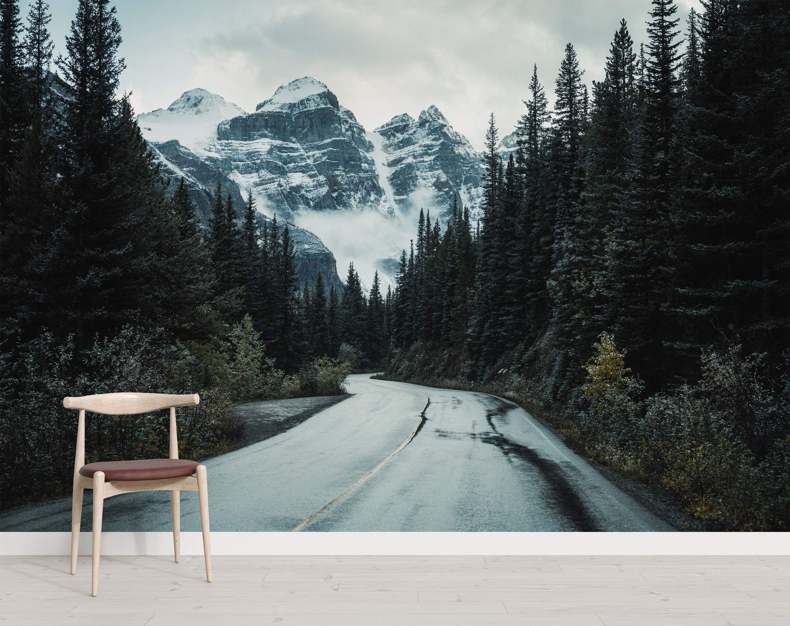 Atmospheric mural of a wet road winding through a pine forest towards snowy mountains under a cloudy, gray sky.