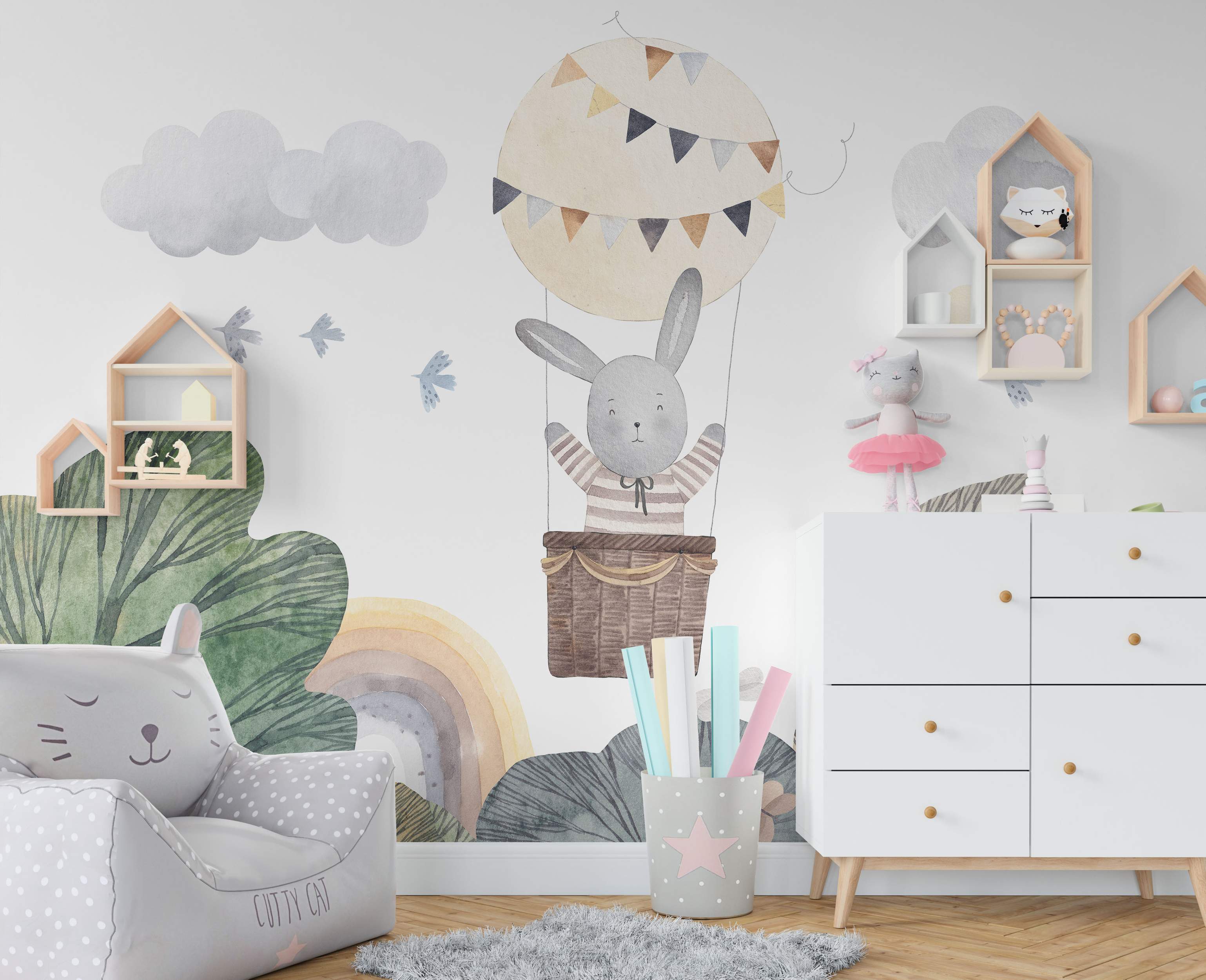 Cute children's wall mural with a watercolor rabbit in a hot air balloon, clouds, birds, rainbow, and hills in muted colors.