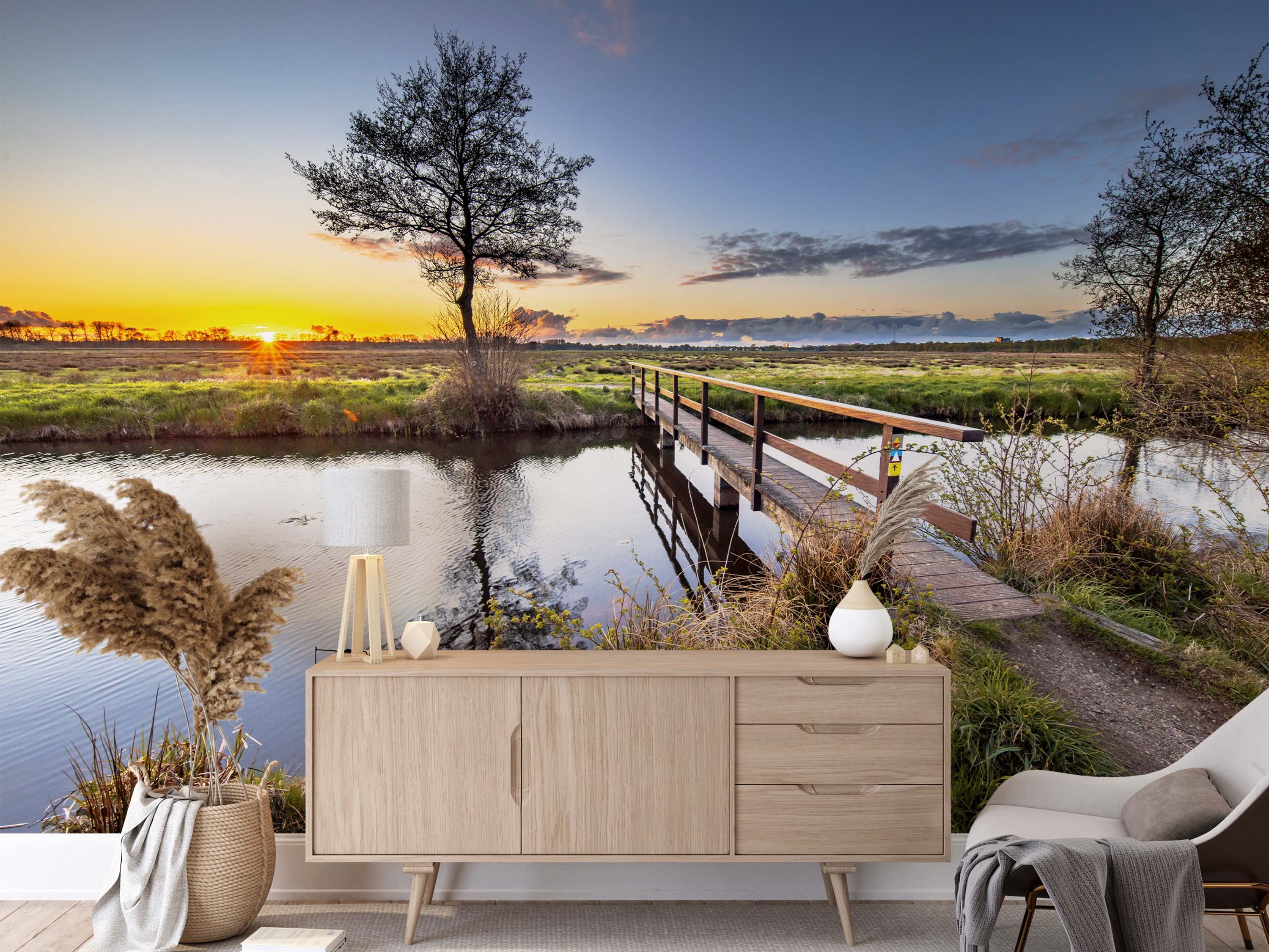 A tranquil landscape mural of a wooden footbridge over a river at sunset, with a silhouetted tree against a golden sky.