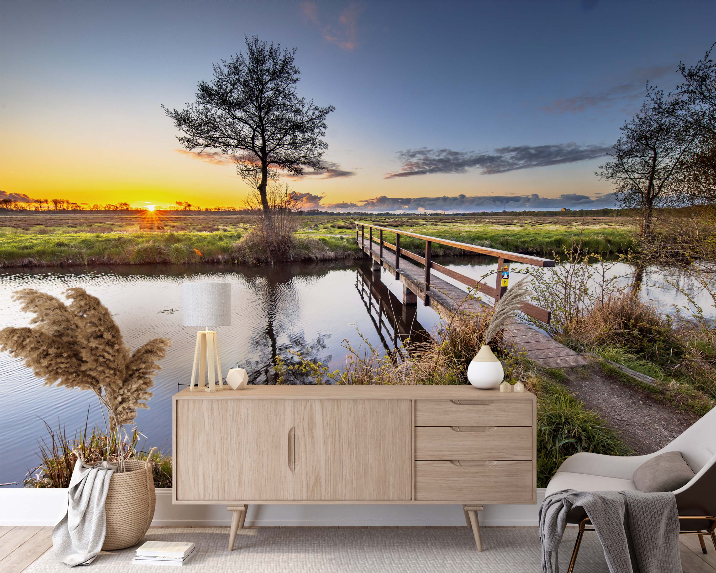 A tranquil landscape mural of a wooden footbridge over a river at sunset, with a silhouetted tree against a golden sky.