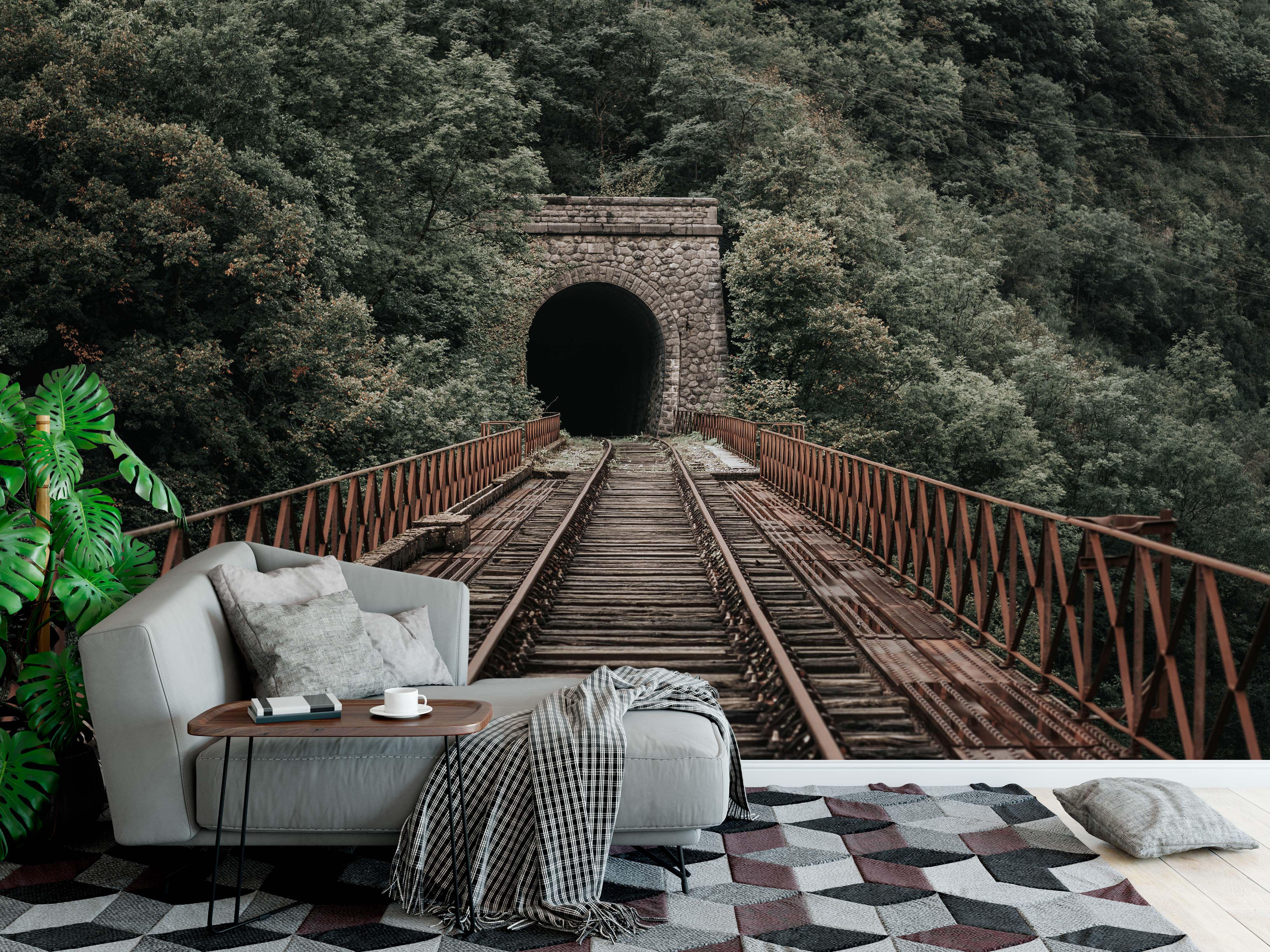 A photographic mural of old, rusty train tracks on a bridge leading into a dark, stone tunnel set in a dense green forest.