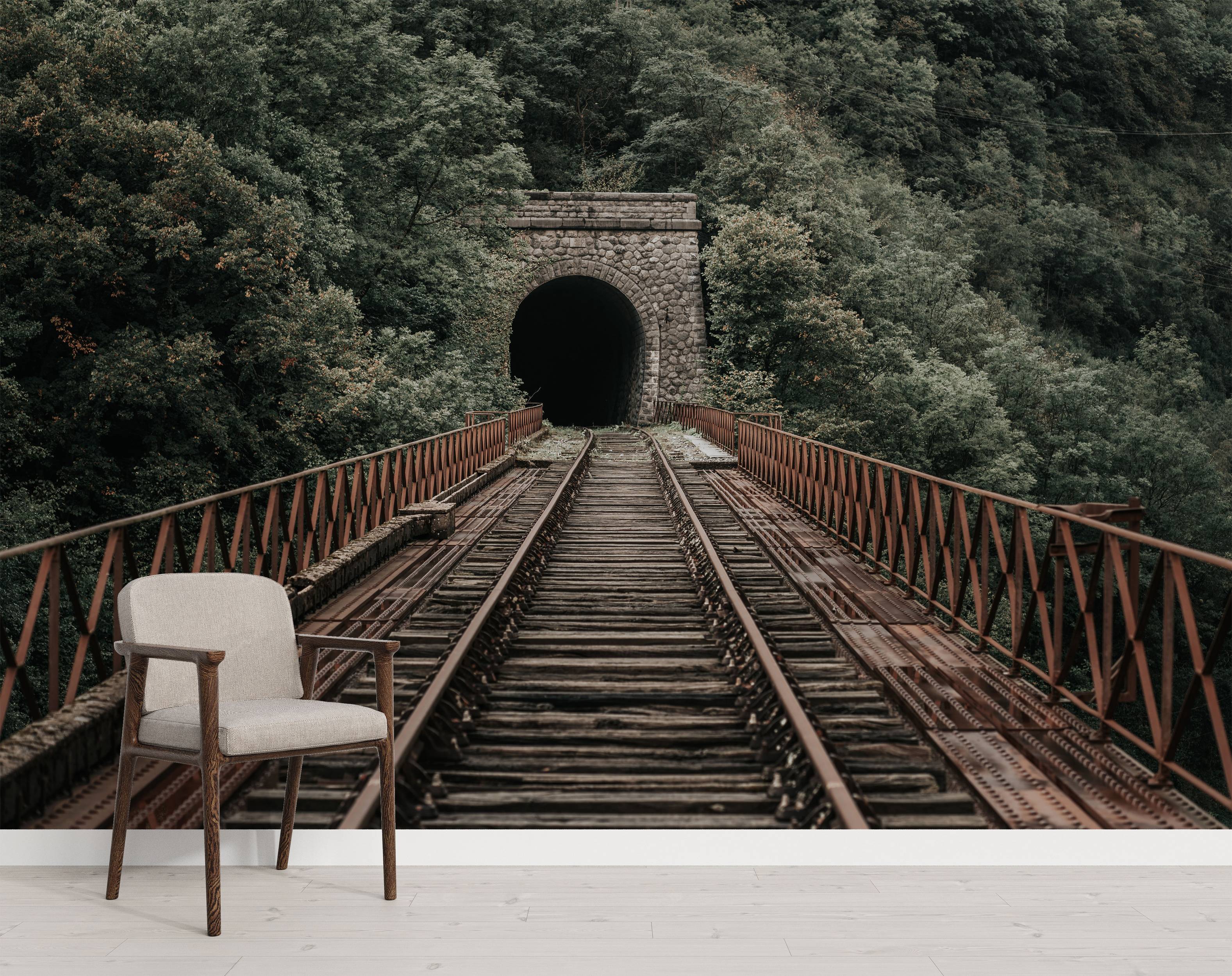 A photographic mural of old train tracks on a rusted bridge leading into a dark stone tunnel surrounded by a dense green forest.
