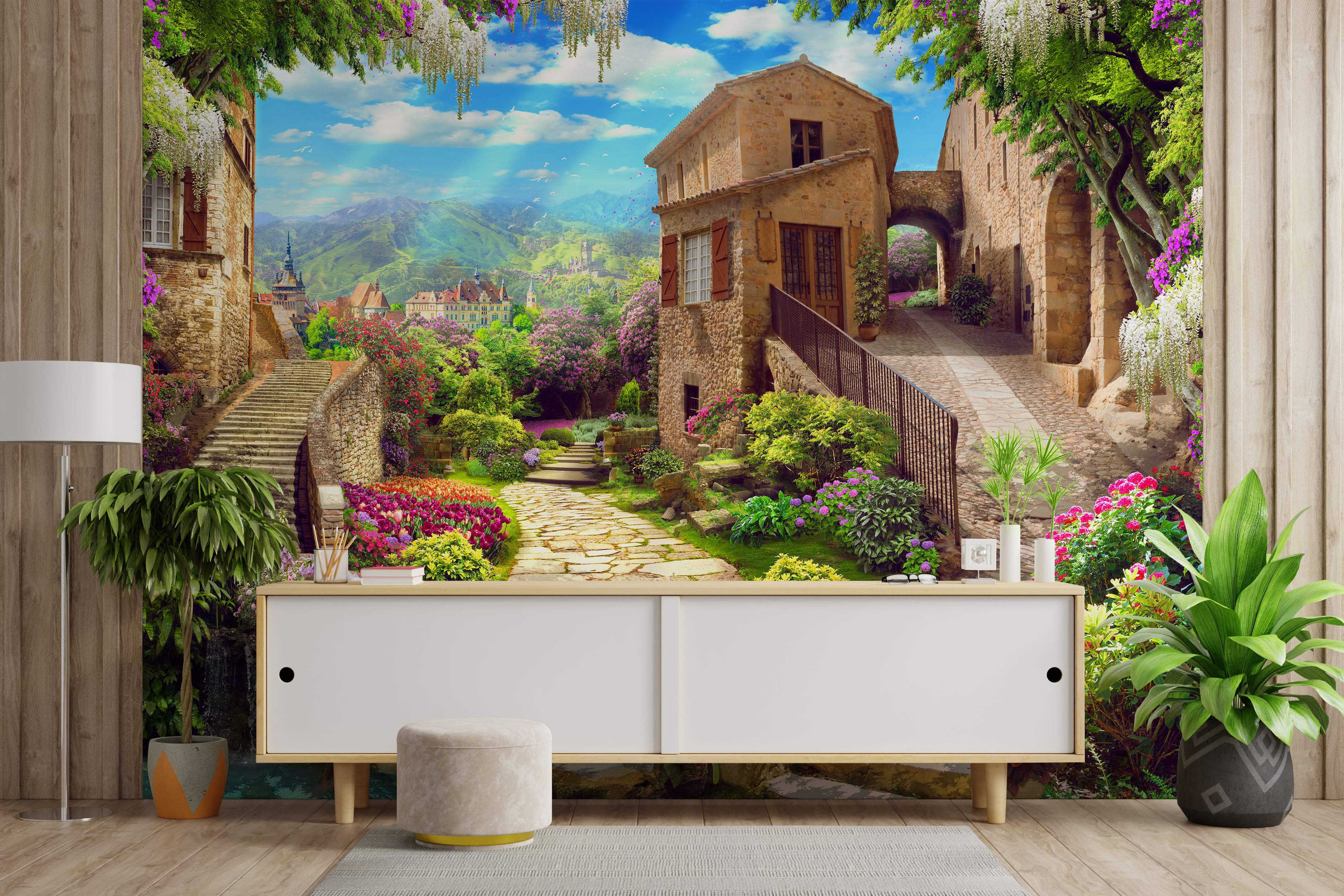 Mural depicting a sunny European village scene with stone buildings, cobblestone paths, stairs, lush gardens, and mountains.
