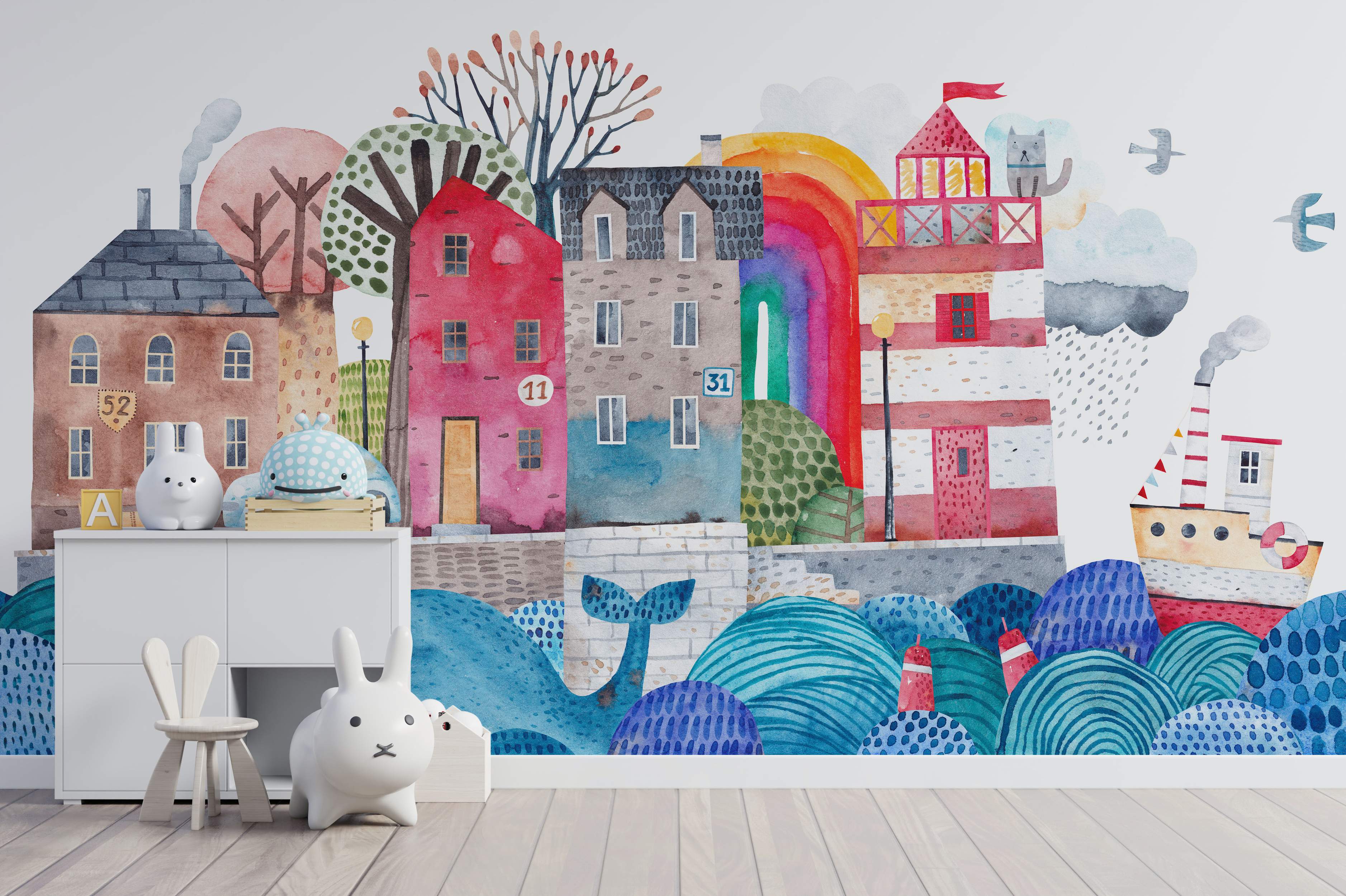 Whimsical watercolor mural depicting a colorful seaside town with houses, a lighthouse, boat, whale, trees, and rainbow.