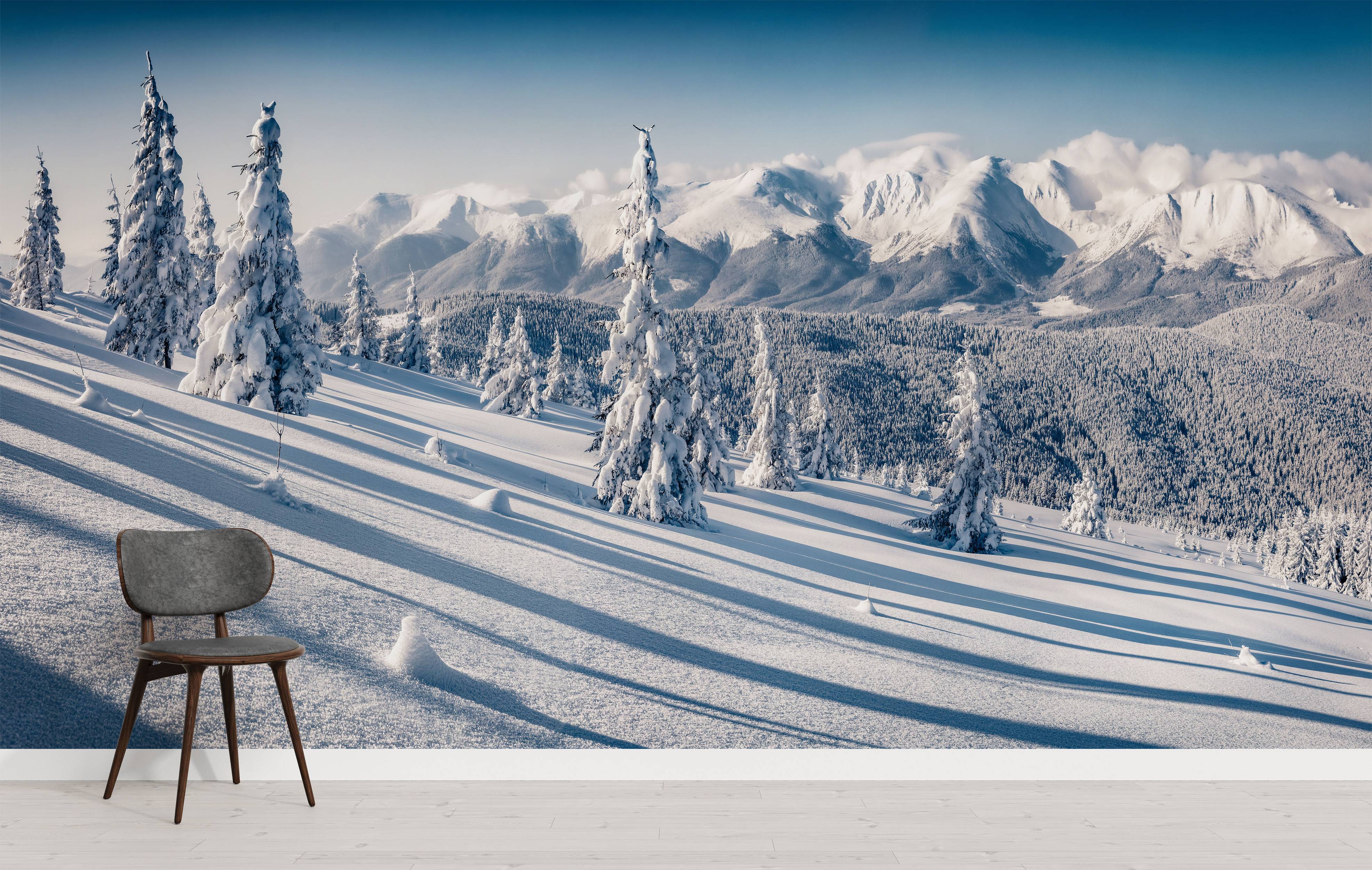 A scenic wall mural of a vast winter mountain range under a clear blue sky, with snow-covered fir trees in the foreground.